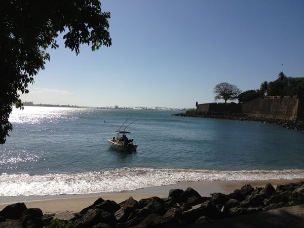 Fishing boat in turquoise waters off a sandy beach with historic fort ruins visible on the coastline. — Plazuela de Isabel I de Castilla, San Juan