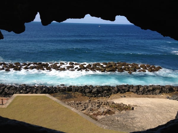 Coastal view framed by a cave opening showing rocky breakwater, turquoise waters, and distant cliffs near Palo Seco… — San Juan