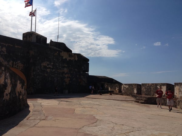 Courtyard of a historic stone fortress with Puerto Rican flags, overlooking the coast under partly cloudy skies. — Faro del Castillo San Felipe del Morro, San Juan