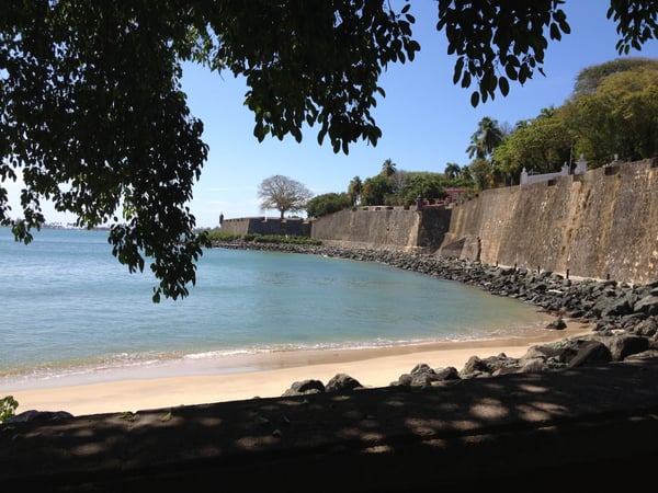 Sandy beach with turquoise waters framed by tree branches, adjacent to colonial-era fortification walls in Old San Juan. — Plazuela de Isabel I de Castilla, San Juan
