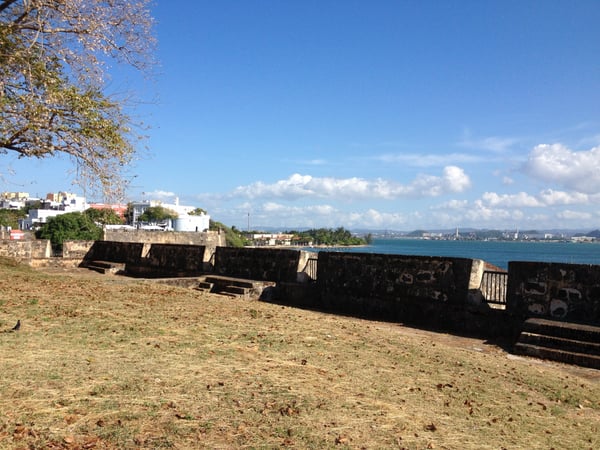 Historic fortification wall on a sandy beach overlooking a turquoise bay with coastal town buildings visible across the… — Viejo San Juan, San Juan