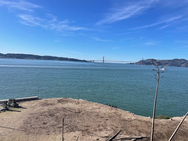 Suspension bridge spanning a bay viewed from a sandy beach with hills and a vessel visible across the water. — Alcatraz Island, San Francisco