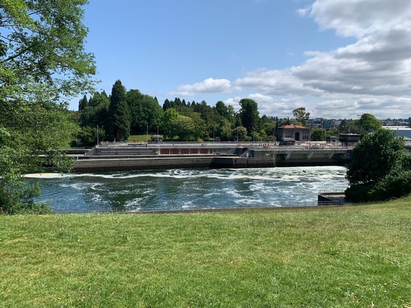Riverfront park with grassy lawn overlooking a flowing river and industrial waterfront structures under partly cloudy… — Ballard, Seattle