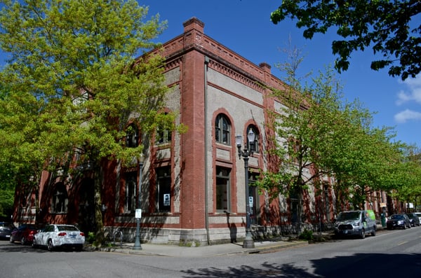 Red brick Romanesque Revival building with stone trim and arched windows, framed by spring foliage on a street corner. — Pearl District, Portland
