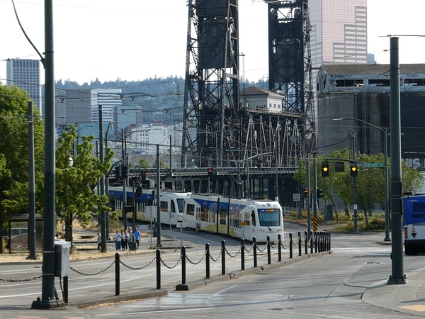 Light rail train passing through downtown Portland with industrial bridges and skyline in the background.