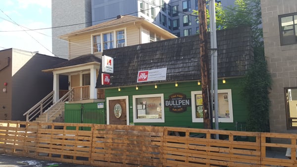 Historic storefront with green facade housing Bullpen restaurant and illy coffee shop, framed by wooden fence and… — Goose Hollow, Portland