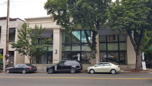 Modern storefront with large windows and mature trees lining a downtown street with parked vehicles. — Goose Hollow, Portland