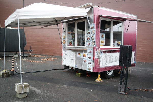 A pink and white Korean food cart with menu boards and decorative plates, set up under a white canopy in a parking lot. — Northwest District, Portland