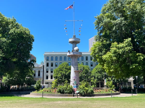 Iconic mid-century tower monument with cross and flags in urban park surrounded by trees and historic buildings. — Tom McCall Waterfront Park, Portland