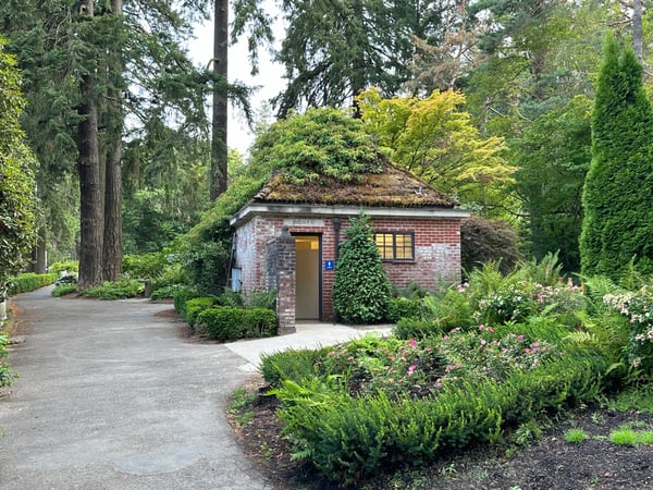 Historic brick restroom building with living roof surrounded by lush gardens and towering conifers in a forest park… — International Rose Test Garden, Portland