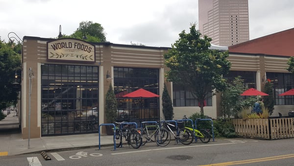 World Foods restaurant storefront with outdoor seating under red umbrellas and bike parking in an urban setting. — Pearl District, Portland