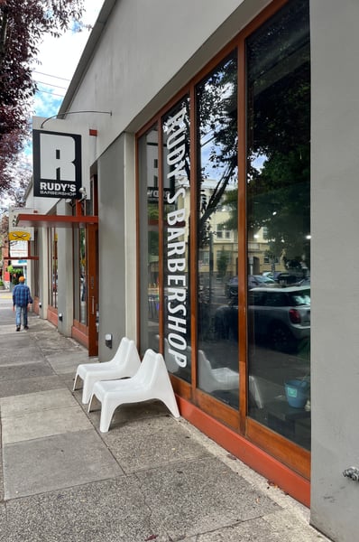 Rudy's Barbershop storefront on a tree-lined urban street with white chairs outside and large windows displaying the… — Northwest District, Portland