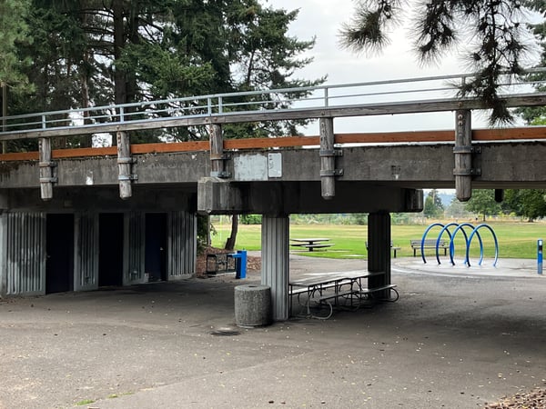 Public park shelter with picnic tables, bike racks, and open green field beyond, framed by concrete overpass and tall… — Portland