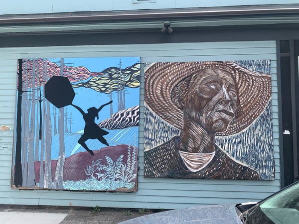 Two large-scale murals depicting a silhouetted figure and a portrait in woodcut style on a building facade in Portland…