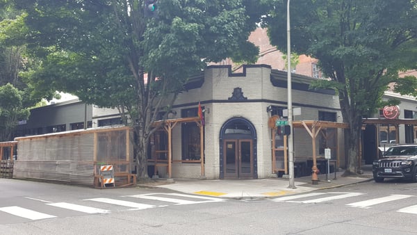 Historic brick building with distinctive curved corner and wooden pergola structures on a tree-lined urban street. — Pearl District, Portland